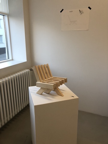Lágur tréstóll með rimlum á sýningarstöpli við glugga.